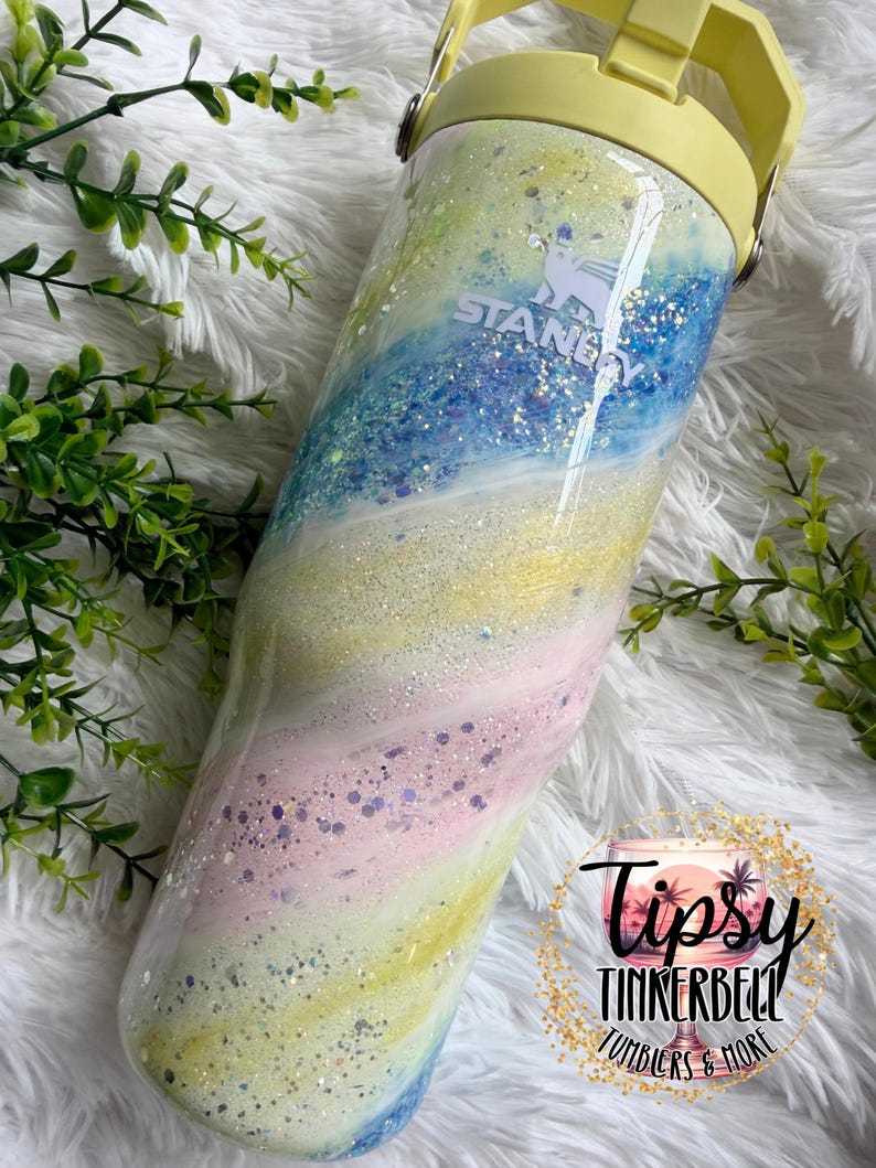May include: A Stanley tumbler with a pastel swirl design in shades of yellow, blue, pink, and white, with glitter accents. The tumbler has a yellow handle and lid. The text "Stanley" is visible on the side. The bottom right corner has the text "Tipsy Tinkerbell Tumblers & More".