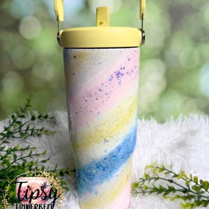 May include: A pastel-colored tumbler with a yellow lid and handle. The tumbler features a swirling design of pink, blue, and yellow with glitter accents. The tumbler has a straw and is designed to keep drinks cold. The logo "Tipsy Tinkerbell Tumblers & More" is visible.
