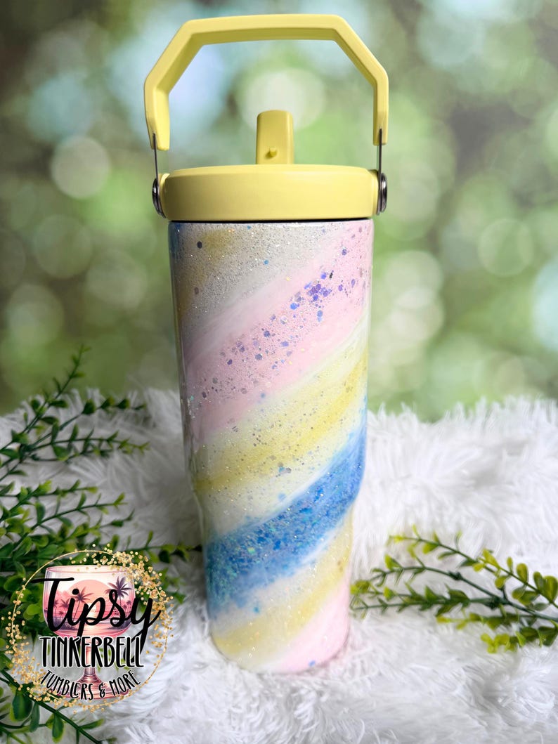 May include: A pastel-colored tumbler with a yellow lid and handle. The tumbler features a swirling design of pink, blue, and yellow with glitter accents. The tumbler has a straw and is designed to keep drinks cold. The logo "Tipsy Tinkerbell Tumblers & More" is visible.