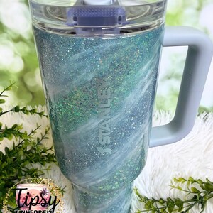 May include: A large, insulated tumbler with a light blue and white glitter swirl design. The tumbler has a clear lid, a gray handle, and the "Stanley" logo. The tumbler is decorated with the text "Tipsy Tinkerbell Tumblers & More".