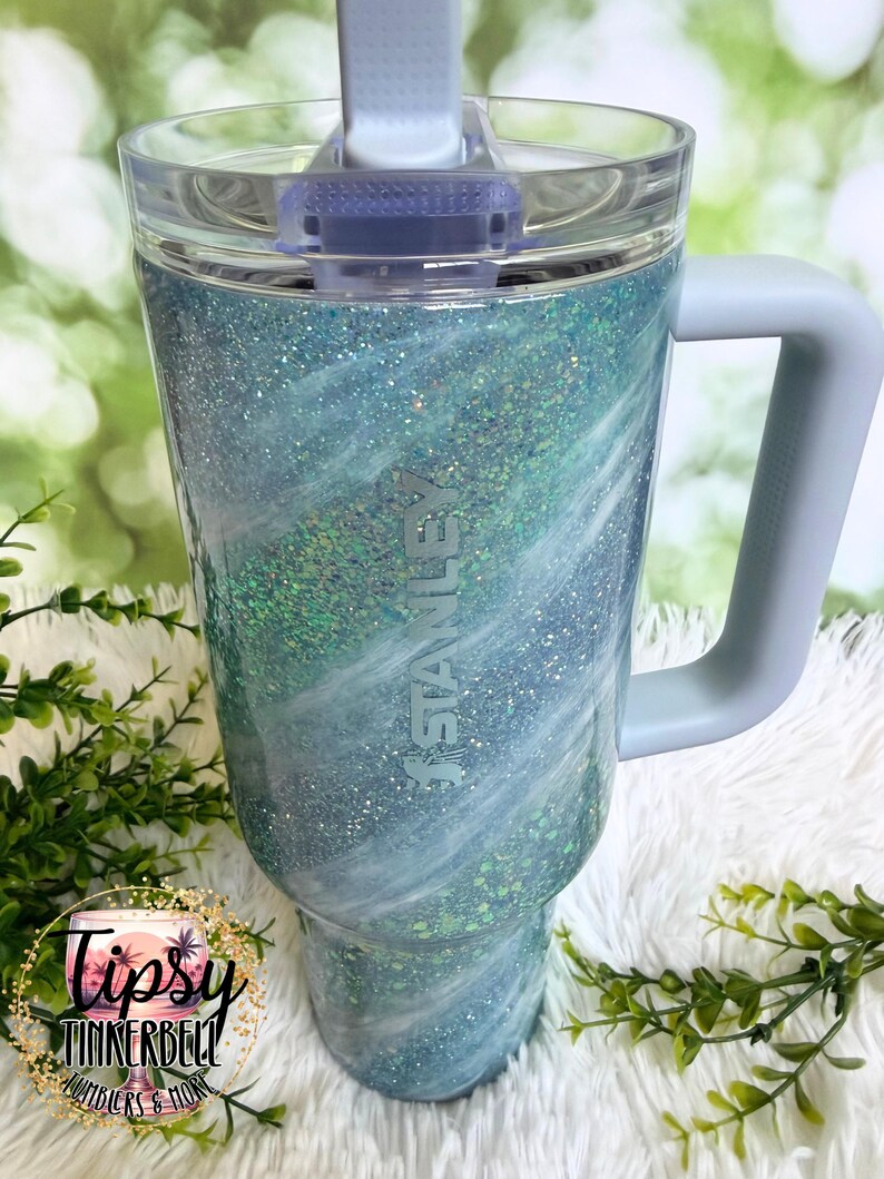 May include: A large, insulated tumbler with a light blue and white glitter swirl design. The tumbler has a clear lid, a gray handle, and the "Stanley" logo. The tumbler is decorated with the text "Tipsy Tinkerbell Tumblers & More".