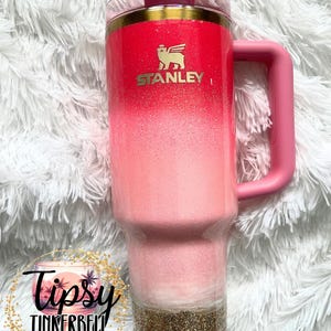 May include: A Stanley tumbler with a pink ombre design, gold accents, and a pink handle. The tumbler features the Stanley logo in gold and a gold glitter base. The tumbler has a straw and a lid. The tumbler is a drinkware item.