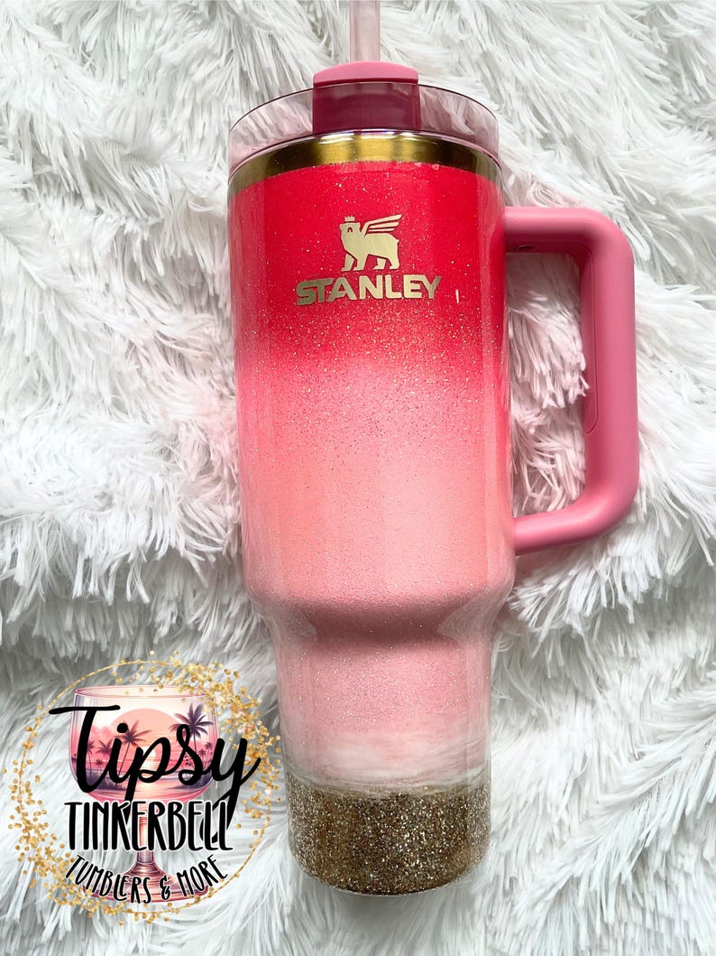 May include: A Stanley tumbler with a pink ombre design, gold accents, and a pink handle. The tumbler features the Stanley logo in gold and a gold glitter base. The tumbler has a straw and a lid. The tumbler is a drinkware item.