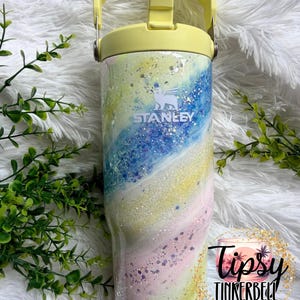 May include: A Stanley tumbler with a pastel swirl design in shades of blue, yellow, pink, and white, accented with glitter. The tumbler has a yellow handle and lid. The Stanley logo is visible on the front.