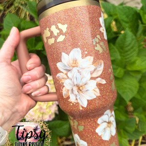 May include: A copper-coloured glitter tumbler with a handle and a dark lid. The surface features a pattern of white floral illustrations and gold leopard print spots. The tumbler has a smooth finish and a cylindrical shape with a tapered base.