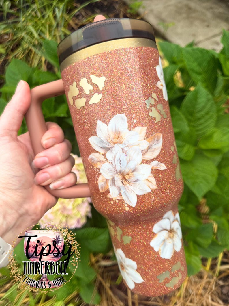 May include: A copper-coloured glitter tumbler with a handle and a dark lid. The surface features a pattern of white floral illustrations and gold leopard print spots. The tumbler has a smooth finish and a cylindrical shape with a tapered base.