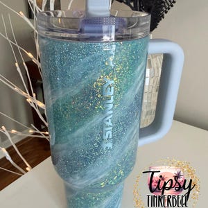 May include: A large, glittery tumbler with a light blue handle and lid. The tumbler has a swirling design of blue, white, and green glitter. The word "STANLEY" is printed on the side. The tumbler is a product of Tipsy Tinkerbell Tumblers & More.