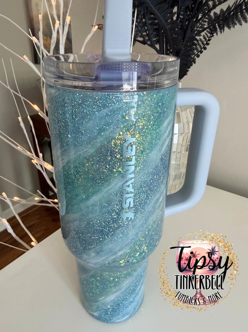 May include: A large, glittery tumbler with a light blue handle and lid. The tumbler has a swirling design of blue, white, and green glitter. The word "STANLEY" is printed on the side. The tumbler is a product of Tipsy Tinkerbell Tumblers & More.