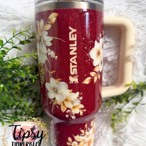 Puede incluir: Un vaso Stanley rojo brillante con un dise&ntilde;o floral. El vaso tiene un asa color crema y el logotipo de Stanley. El dise&ntilde;o floral presenta flores blancas y amarillas y hojas verdes. El vaso est&aacute; etiquetado como "Tipsy Tinkerbell Tumblers & More."