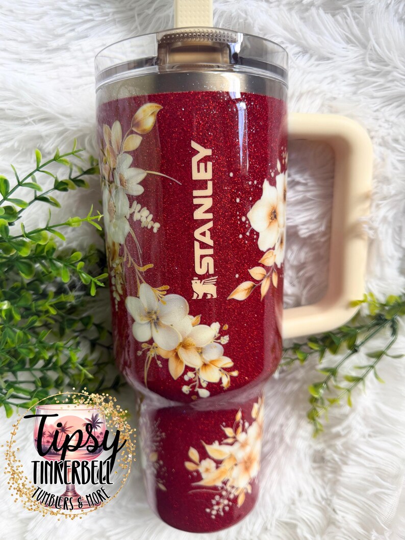 Puede incluir: Un vaso Stanley rojo brillante con un dise&ntilde;o floral. El vaso tiene un asa color crema y el logotipo de Stanley. El dise&ntilde;o floral presenta flores blancas y amarillas y hojas verdes. El vaso est&aacute; etiquetado como "Tipsy Tinkerbell Tumblers & More."