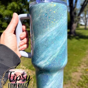 May include: A large, insulated tumbler with a handle, featuring a swirling blue and white design with glitter accents. The tumbler has a clear lid and a gray handle. The logo "Tipsy Tinkerbell Tumblers & More" is visible.