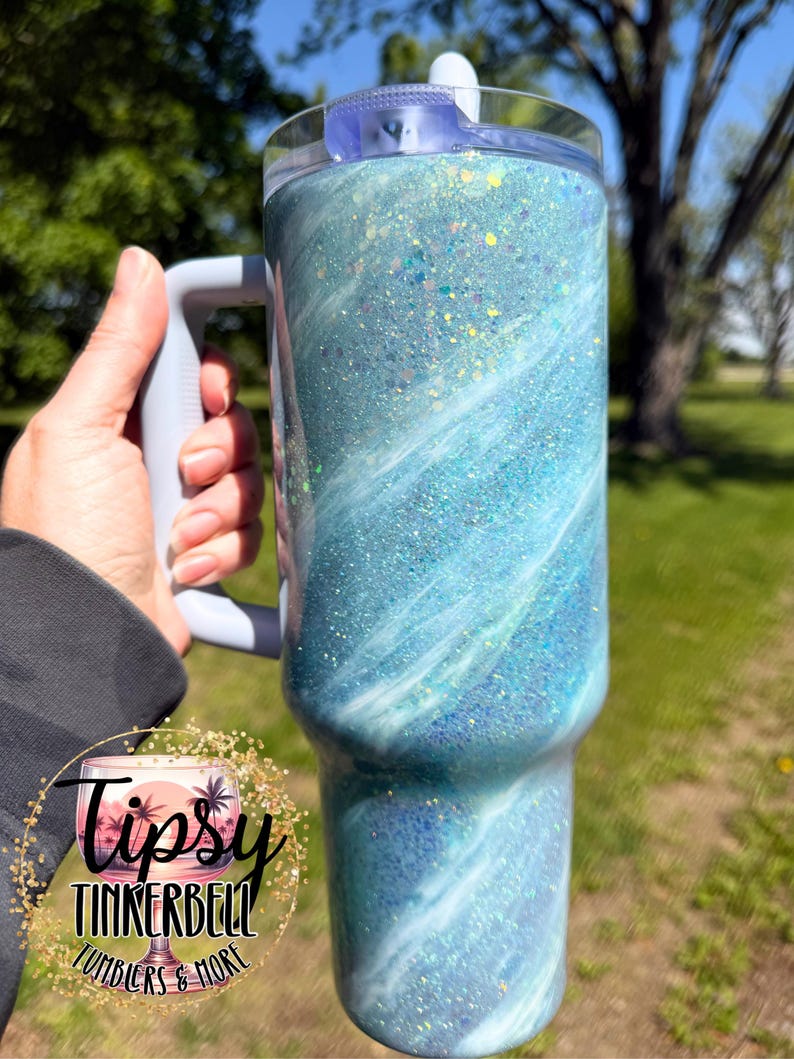 May include: A large, insulated tumbler with a handle, featuring a swirling blue and white design with glitter accents. The tumbler has a clear lid and a gray handle. The logo "Tipsy Tinkerbell Tumblers & More" is visible.