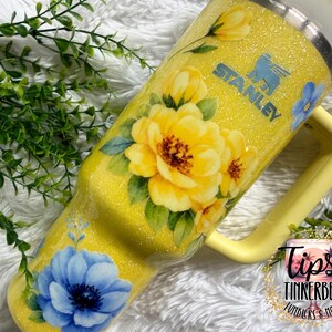 May include: A yellow Stanley tumbler with a floral design featuring large yellow and blue flowers. The tumbler has a handle and a glittery finish. The Stanley logo is visible on the side.