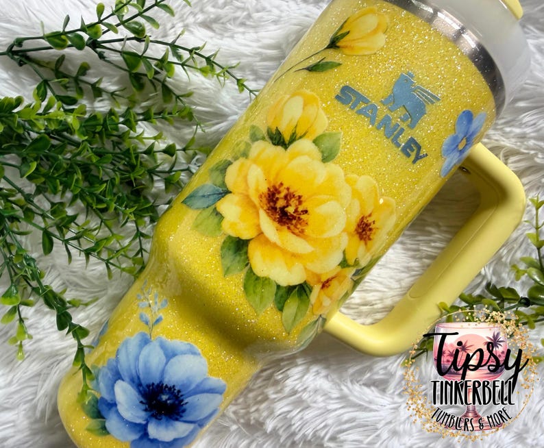 May include: A yellow Stanley tumbler with a floral design featuring large yellow and blue flowers. The tumbler has a handle and a glittery finish. The Stanley logo is visible on the side.