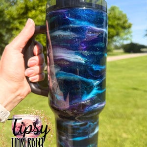 May include: A large, insulated tumbler with a black handle and lid. The tumbler features a swirling design of blues, purples, and white, with a glittery finish. The tumbler has the text "Tipsy Tinkerbell Tumblers & More" on the bottom.
