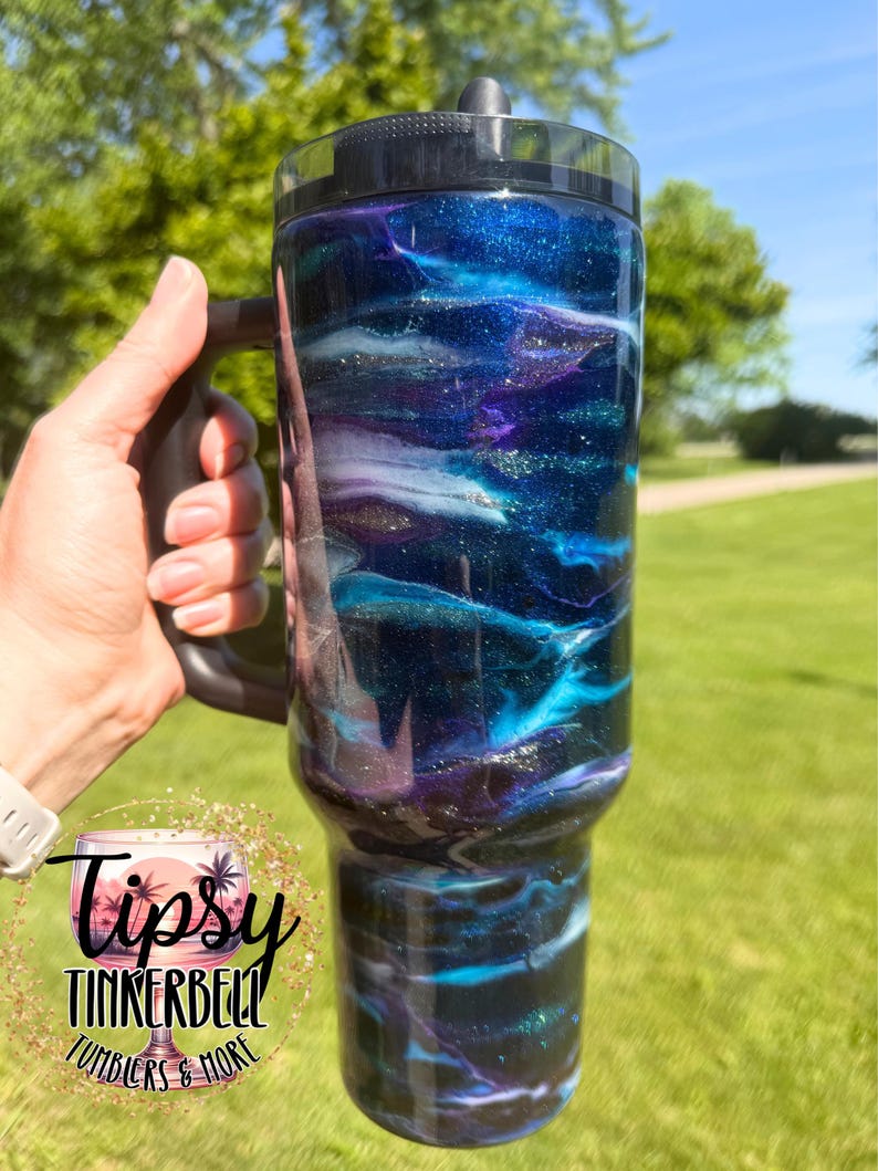 May include: A large, insulated tumbler with a black handle and lid. The tumbler features a swirling design of blues, purples, and white, with a glittery finish. The tumbler has the text "Tipsy Tinkerbell Tumblers & More" on the bottom.