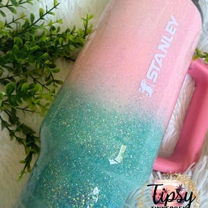 May include: A Stanley tumbler featuring a glittery ombre finish that transitions from light pink at the top to teal at the bottom. The cup includes a pink handle, a clear lid, and the white Stanley logo printed on the upper pink section.