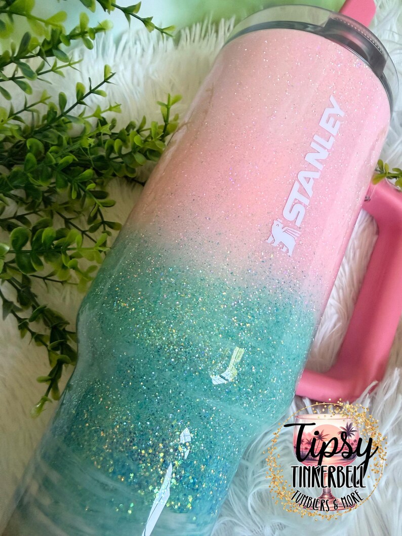 May include: A Stanley tumbler featuring a glittery ombre finish that transitions from light pink at the top to teal at the bottom. The cup includes a pink handle, a clear lid, and the white Stanley logo printed on the upper pink section.