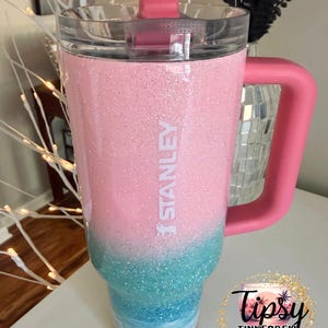 May include: A Stanley tumbler featuring a pink and blue glitter ombre finish. The cup has a clear lid, a pink handle, and a pink straw. The word STANLEY is printed vertically in white on the upper pink section of the glitter-coated exterior.