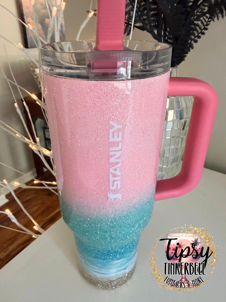 May include: A Stanley tumbler featuring a pink and blue glitter ombre finish. The cup has a clear lid, a pink handle, and a pink straw. The word STANLEY is printed vertically in white on the upper pink section of the glitter-coated exterior.
