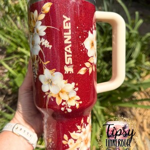 Puede incluir: Un vaso Stanley rojo brillante con un dise&ntilde;o floral. El vaso tiene un asa color crema y una tapa transparente. El dise&ntilde;o floral presenta flores blancas y amarillas con hojas verdes. El logotipo de Stanley es visible.