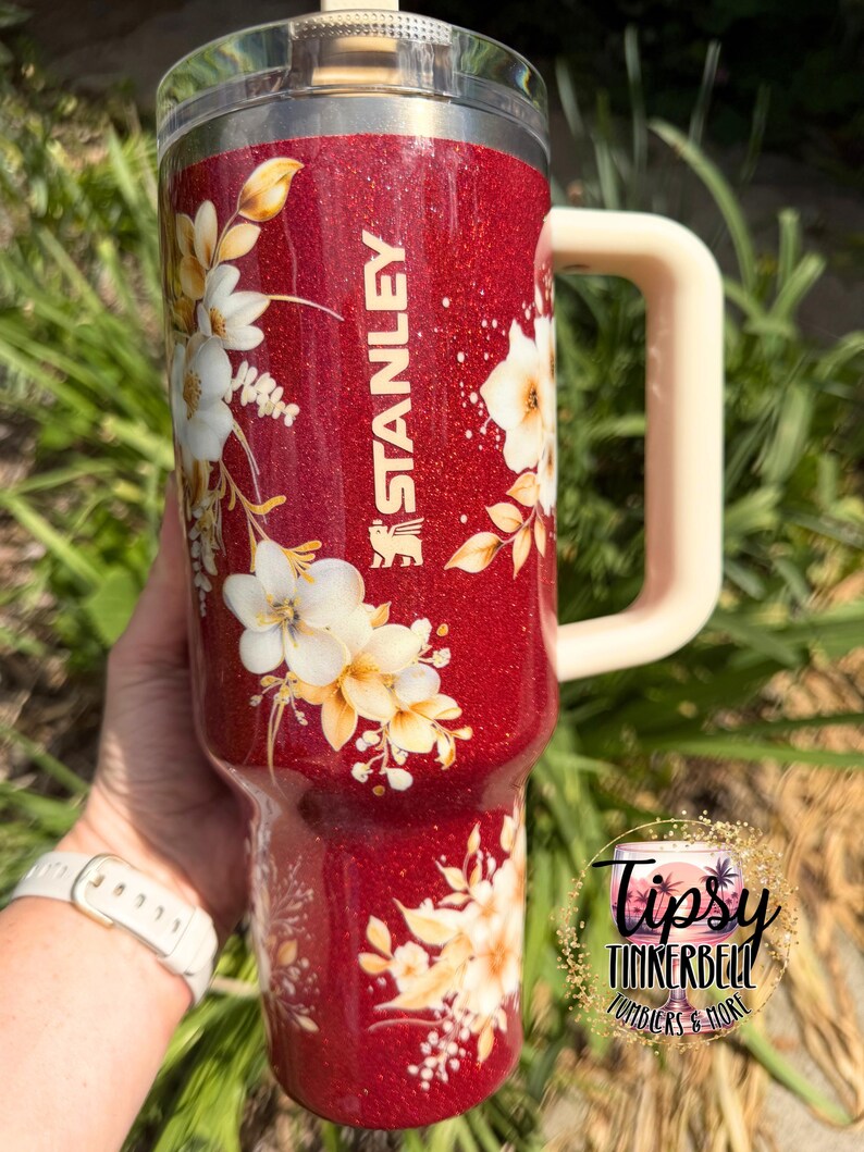 Puede incluir: Un vaso Stanley rojo brillante con un dise&ntilde;o floral. El vaso tiene un asa color crema y una tapa transparente. El dise&ntilde;o floral presenta flores blancas y amarillas con hojas verdes. El logotipo de Stanley es visible.
