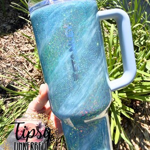May include: A large, blue glitter tumbler with a light blue handle and lid. The tumbler has a swirling design with shades of blue and green. The text "TUMBLERS & MORE" is visible on the bottom left.