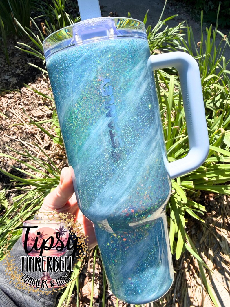 May include: A large, blue glitter tumbler with a light blue handle and lid. The tumbler has a swirling design with shades of blue and green. The text "TUMBLERS & MORE" is visible on the bottom left.
