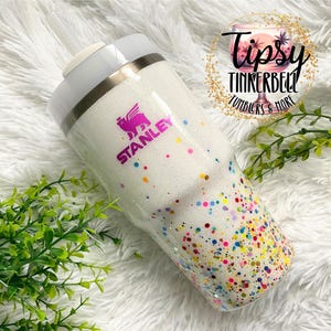 Puede incluir: Un vaso Stanley blanco con acabado brillante y puntos de confeti coloridos. El vaso tiene una tapa blanca y una pajita. El logotipo de Stanley es visible en rosa. El texto "Tipsy Tinkerbell Tumblers & More" tambi&eacute;n es visible.