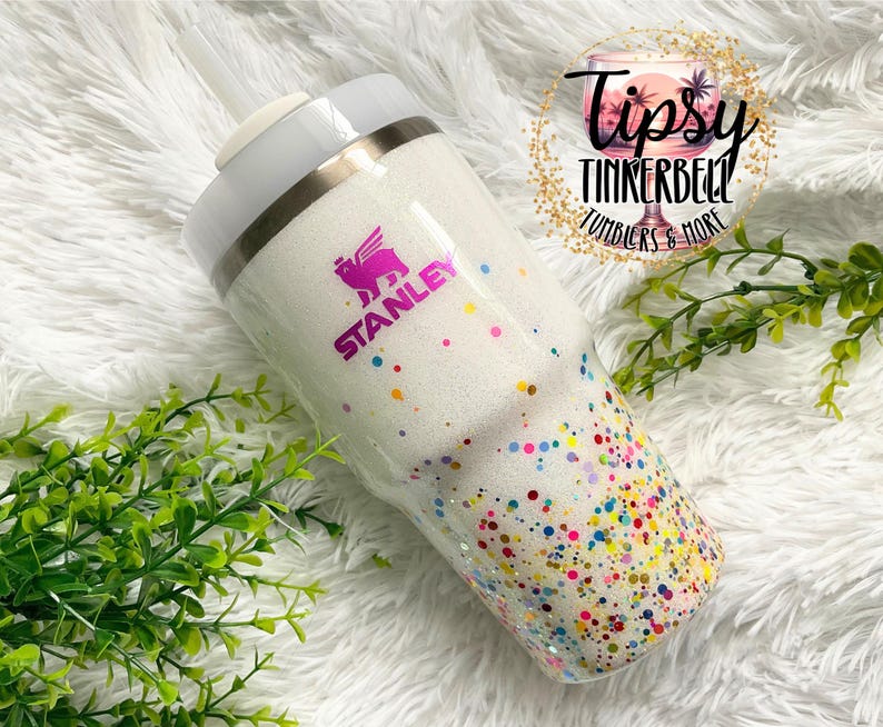 Puede incluir: Un vaso Stanley blanco con acabado brillante y puntos de confeti coloridos. El vaso tiene una tapa blanca y una pajita. El logotipo de Stanley es visible en rosa. El texto "Tipsy Tinkerbell Tumblers & More" tambi&eacute;n es visible.