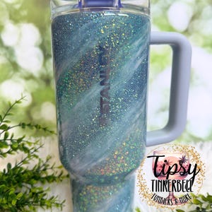 May include: A glittery blue tumbler with a handle and a lid. The tumbler has a swirling design with white accents and the word "STANLEY" printed on the side. The tumbler is approximately 9 inches tall.