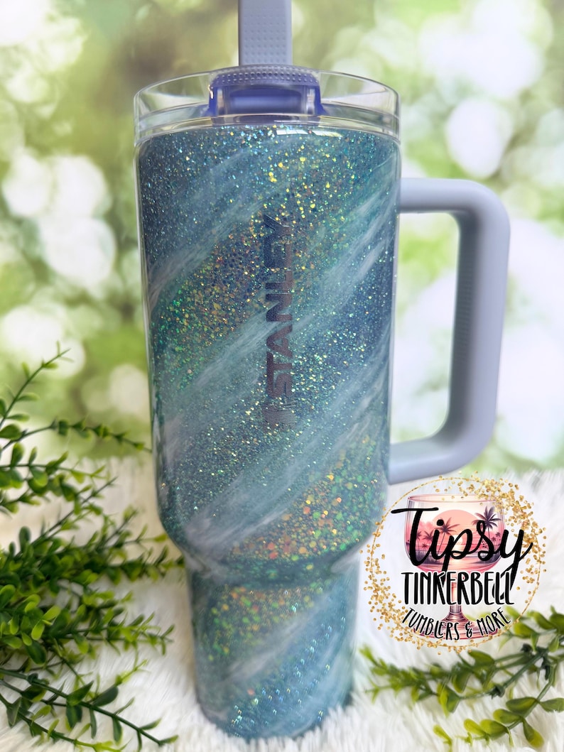 May include: A glittery blue tumbler with a handle and a lid. The tumbler has a swirling design with white accents and the word "STANLEY" printed on the side. The tumbler is approximately 9 inches tall.