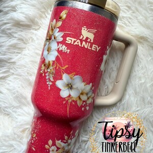 K&ouml;nnte beinhalten: Ein leuchtend pinker Glitzer-Stanley-Becher mit beigem Griff und Deckel. Der Becher zeigt ein wei&szlig;es Blumenmuster mit goldenen Akzenten und das Stanley-Logo in Gold. Ein kreisf&ouml;rmiges Logo mit dem Text Tipsy Tinkerbell Tumblers & More ist zu sehen.