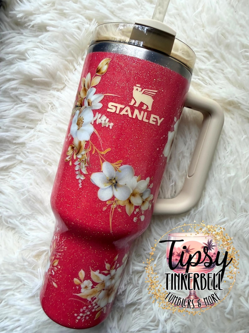K&ouml;nnte beinhalten: Ein leuchtend pinker Glitzer-Stanley-Becher mit beigem Griff und Deckel. Der Becher zeigt ein wei&szlig;es Blumenmuster mit goldenen Akzenten und das Stanley-Logo in Gold. Ein kreisf&ouml;rmiges Logo mit dem Text Tipsy Tinkerbell Tumblers & More ist zu sehen.