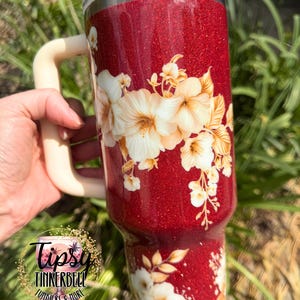 Puede incluir: Un vaso rojo brillante con un asa color crema y un dise&ntilde;o floral. El vaso presenta flores blancas y doradas. El vaso tiene una tapa transparente y el logotipo "Tipsy Tinkerbell Tumblers & More".