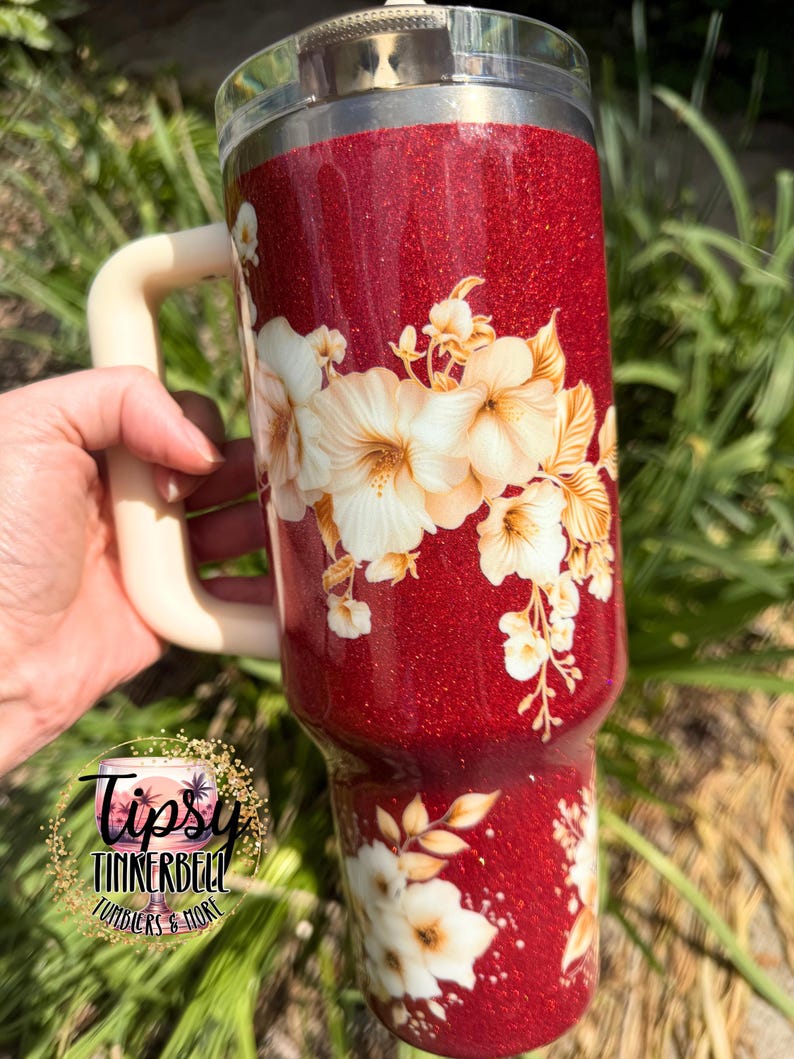 Puede incluir: Un vaso rojo brillante con un asa color crema y un dise&ntilde;o floral. El vaso presenta flores blancas y doradas. El vaso tiene una tapa transparente y el logotipo "Tipsy Tinkerbell Tumblers & More".