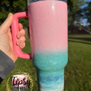 May include: A tall stainless steel tumbler with a handle and a clear lid, featuring a glittery ombre design that transitions from a light pink top to a turquoise middle, ending in a painted beach scene with blue waves and a sandy base.