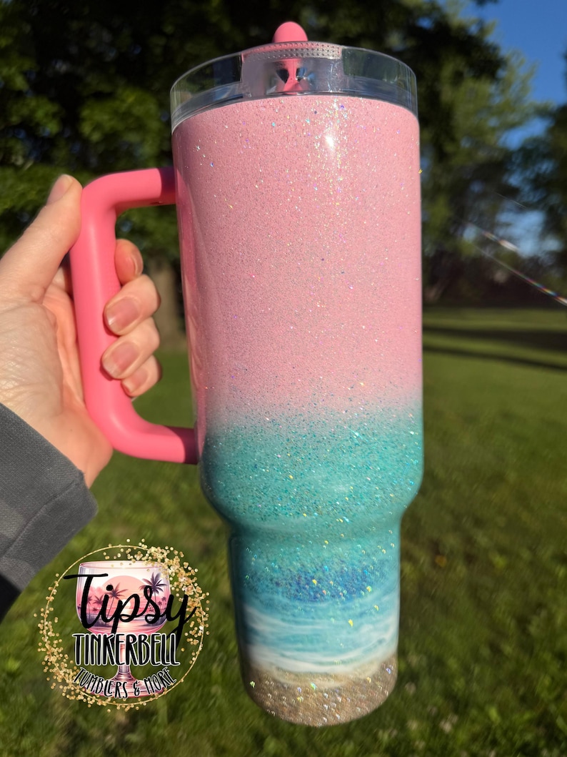 May include: A tall stainless steel tumbler with a handle and a clear lid, featuring a glittery ombre design that transitions from a light pink top to a turquoise middle, ending in a painted beach scene with blue waves and a sandy base.