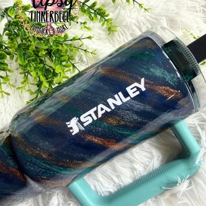 Puede incluir: Un vaso Stanley con asa turquesa y correa negra. El vaso tiene un dise&ntilde;o de purpurina azul marino, turquesa y dorado. La palabra "STANLEY" est&aacute; impresa en blanco en el lateral del vaso.