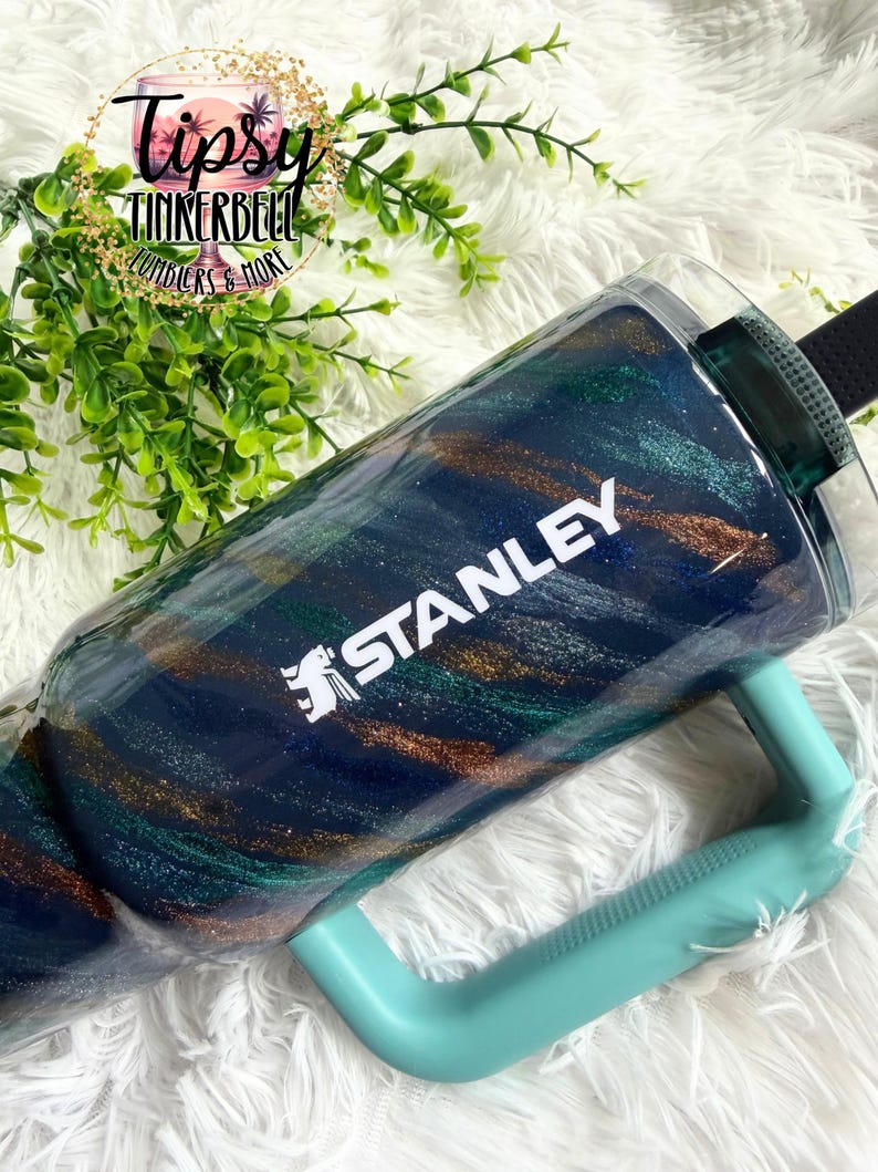 Puede incluir: Un vaso Stanley con asa turquesa y correa negra. El vaso tiene un dise&ntilde;o de purpurina azul marino, turquesa y dorado. La palabra "STANLEY" est&aacute; impresa en blanco en el lateral del vaso.