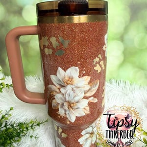 May include: A copper-coloured glitter tumbler with a handle and a dark brown lid featuring a straw. The exterior is decorated with white floral illustrations and gold leopard print accents. A circular logo reads Tipsy Tinkerbell Tumblers & More.