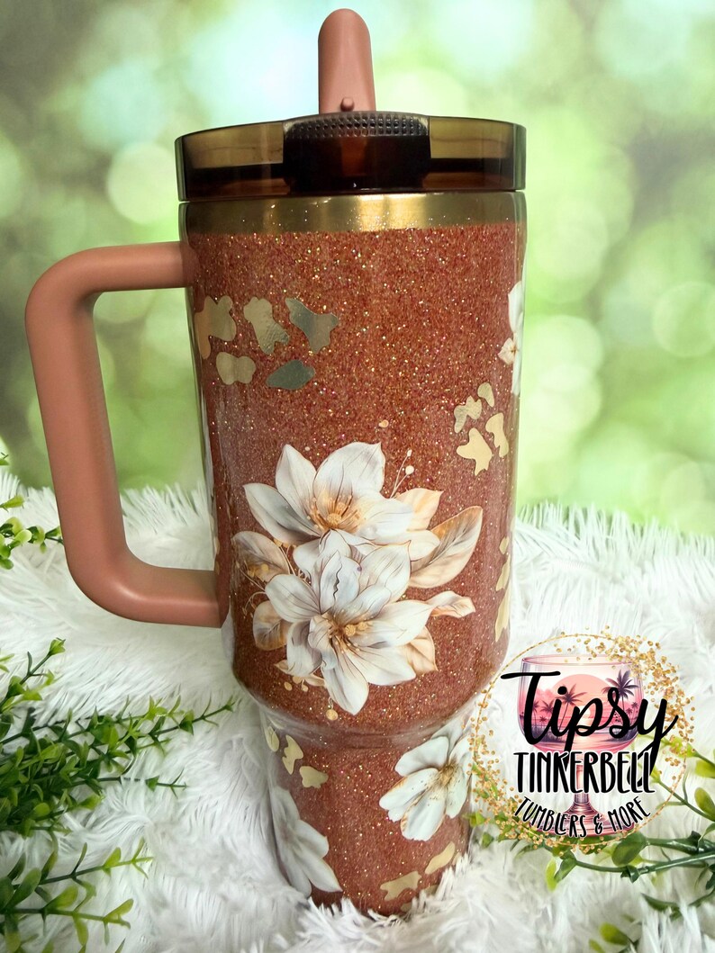 May include: A copper-coloured glitter tumbler with a handle and a dark brown lid featuring a straw. The exterior is decorated with white floral illustrations and gold leopard print accents. A circular logo reads Tipsy Tinkerbell Tumblers & More.