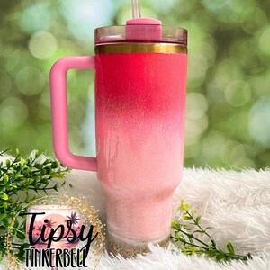 May include: A pink ombre tumbler with a handle and straw. The tumbler features a gradient design, transitioning from a deep pink at the top to a lighter pink at the bottom, with a gold band. The base has a sand-like texture. The tumbler has the text "Tipsy Tinkerbell Tumblers & More".