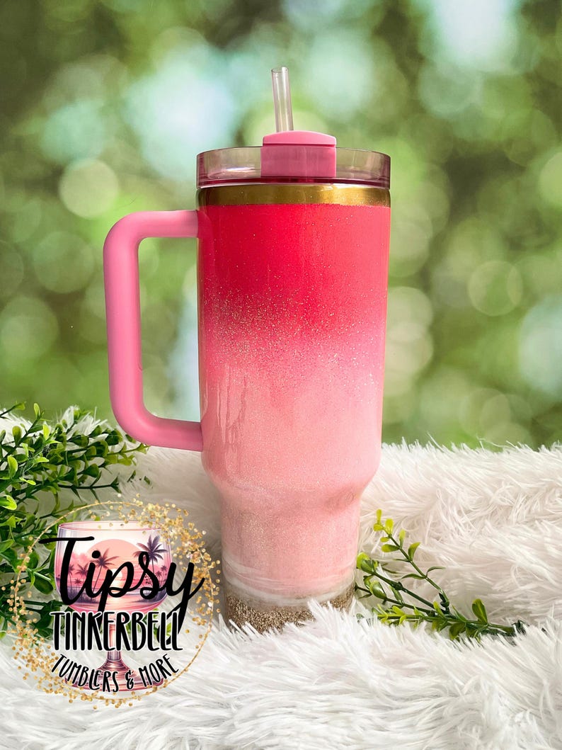 May include: A pink ombre tumbler with a handle and straw. The tumbler features a gradient design, transitioning from a deep pink at the top to a lighter pink at the bottom, with a gold band. The base has a sand-like texture. The tumbler has the text "Tipsy Tinkerbell Tumblers & More".