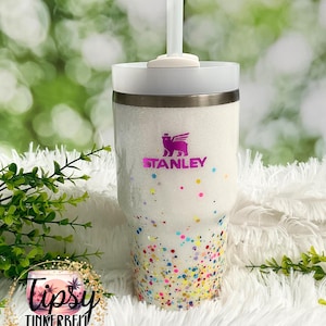 Puede incluir: Un vaso Stanley blanco con acabado brillante y detalles de confeti de colores. El vaso presenta el logotipo de Stanley en morado y el texto "Tipsy Tinkerbell Tumblers & More". Incluye tapa y pajita blancas.