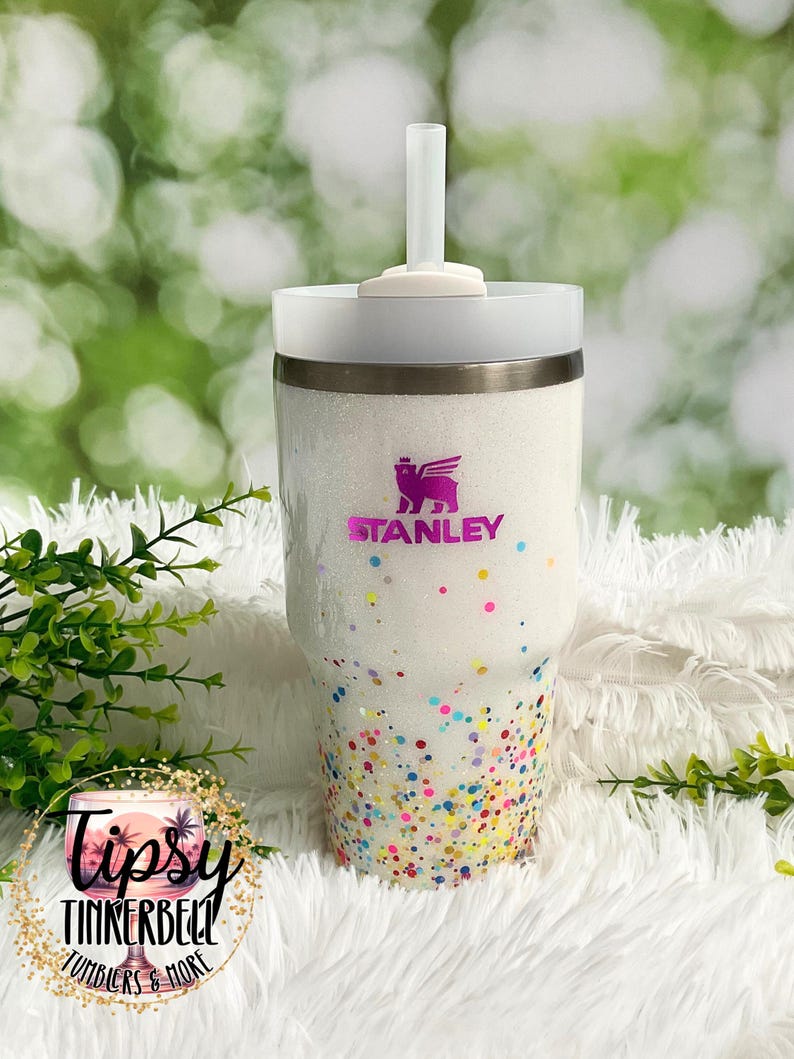 Puede incluir: Un vaso Stanley blanco con acabado brillante y detalles de confeti de colores. El vaso presenta el logotipo de Stanley en morado y el texto "Tipsy Tinkerbell Tumblers & More". Incluye tapa y pajita blancas.