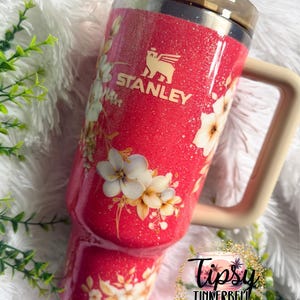 K&ouml;nnte beinhalten: Ein leuchtend pinkfarbener Stanley-Becher mit Glitzer-Finish und einem Blumenmuster aus wei&szlig;en und cremefarbenen Bl&uuml;ten. Der Becher hat einen beigen Henkel, einen dunkel durchscheinenden Deckel und einen Strohhalm. Ein rundes Logo zeigt Tipsy Tinkerbell Tumblers & More.