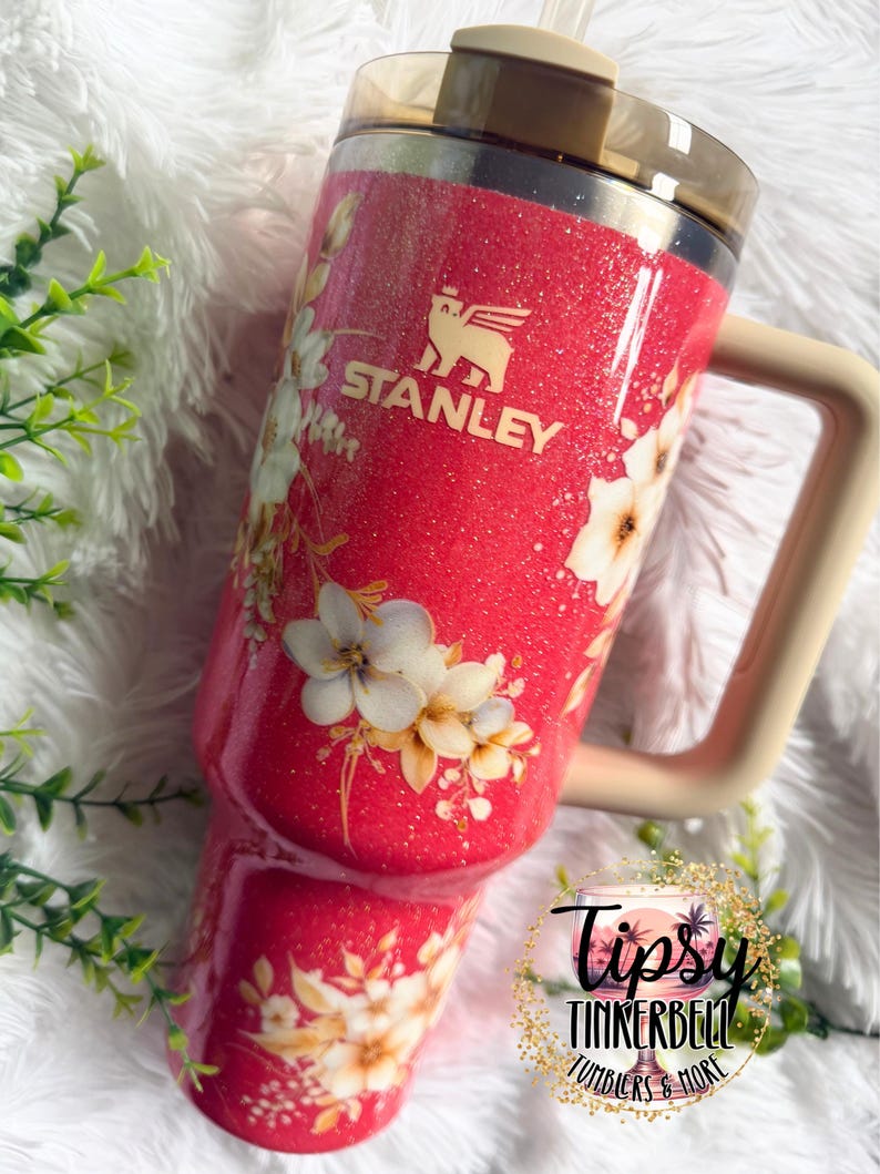 K&ouml;nnte beinhalten: Ein leuchtend pinkfarbener Stanley-Becher mit Glitzer-Finish und einem Blumenmuster aus wei&szlig;en und cremefarbenen Bl&uuml;ten. Der Becher hat einen beigen Henkel, einen dunkel durchscheinenden Deckel und einen Strohhalm. Ein rundes Logo zeigt Tipsy Tinkerbell Tumblers & More.