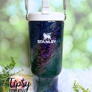 May include: A Stanley tumbler featuring a dark, glittery finish with swirling shades of blue, purple, and green. The cup has a white lid with a built-in handle and straw mechanism. The white Stanley logo is printed on the upper front of the tumbler.