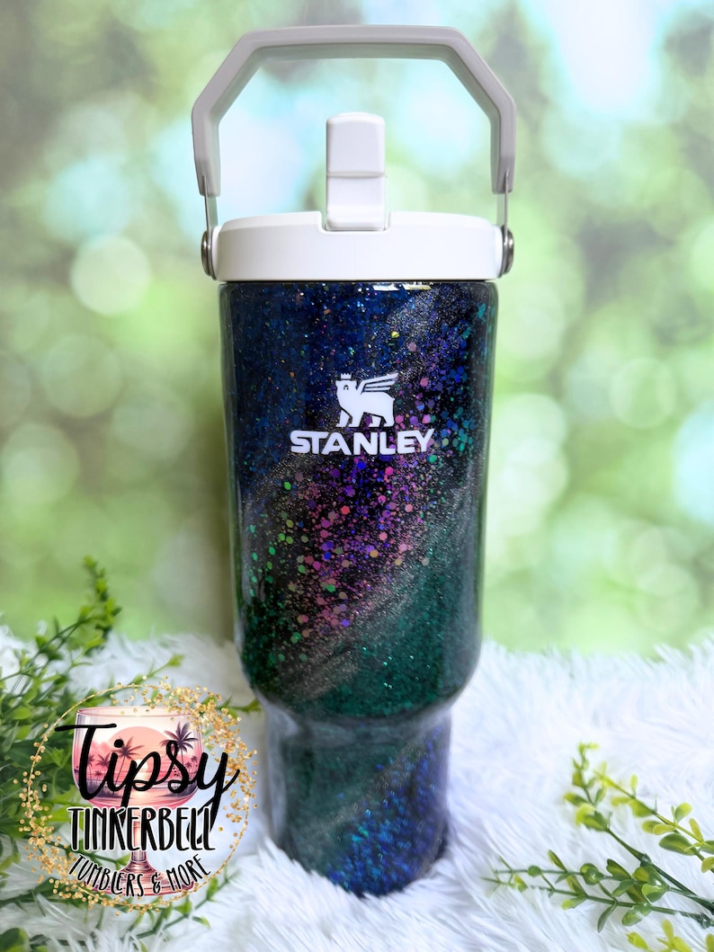 May include: A Stanley tumbler featuring a dark, glittery finish with swirling shades of blue, purple, and green. The cup has a white lid with a built-in handle and straw mechanism. The white Stanley logo is printed on the upper front of the tumbler.